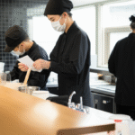 Tiga pekerja dengan seragam hitam dan masker sedang bekerja di dapur komersial yang bersih, salah satunya memegang kertas seperti sedang membaca instruksi atau resep.