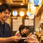 Seorang staf pria muda tersenyum ramah saat melayani pelanggan di sebuah Izakaya yang ramai di Tokyo.