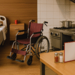 Perbedaan jalur TITP dan Tokutei Ginou bidang caregiving dan restoran, digambarkan secara visual melalui sudut ruangan yang terbagi dua: sisi kiri menunjukkan suasana ruang perawatan lansia dengan tempat tidur rumah sakit dan kursi roda beraksen merah, sedangkan sisi kanan menggambarkan dapur restoran profesional lengkap dengan perlengkapan memasak, menu beraksen merah, serta detail kontras modern dan fungsional. Ilustrasi tanpa kehadiran manusia, menonjolkan perbedaan atmosfer kerja antara dua jalur ini secara realistik dan unik.