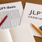 Foto realistik meja belajar berisi lembar ujian JFT Basic dengan pulpen merah, buku JLPT dengan pensil dan penghapus abu-abu, serta buku catatan terbuka. Visual ini menggambarkan ujian JFT Basic vs JLPT perbedaan strategi persiapan, dengan nuansa natural, aksen merah, dan tanpa kehadiran manusia.
