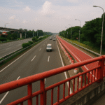 Foto realistik jalan tol dari Karawang menuju lokasi ujian, diambil dari jembatan dengan sudut unik. Menampilkan jalur kendaraan ramai lancar, jalur khusus berwarna merah, serta pagar jembatan merah mencolok. Visual ini menggambarkan rute akses ujian dari Karawang ke lokasi dengan nuansa profesional, modern, dan dinamis.
