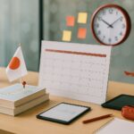 Roadmap belajar bahasa Jepang enam bulan untuk SSW dari Karawang, set meja studi modern tanpa manusia, aksen merah, suasana sekolah bahasa yang profesional.